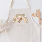 Kinderschürze mit floralem Boho-Regenbogen und personalisiert mit einem Namen mit pastellgrünen Großbuchstaben